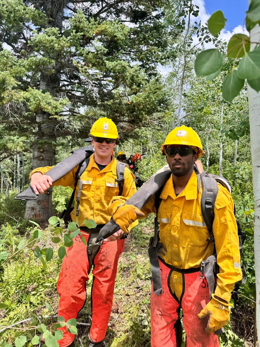 Two men hold grey hoses over their shoulders while wearing orange pants, yellow tops and yellow hard hats. They're standing among trees.