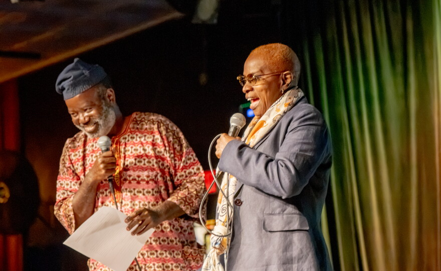 A man and woman stand on stage holding microphones and laughing