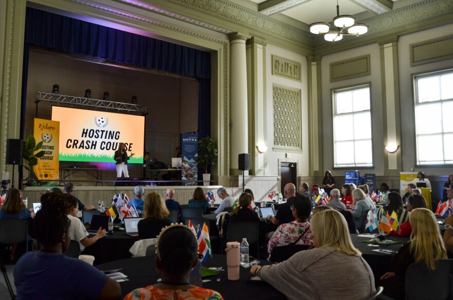 People sit around a table. A speaker on the stage is talking into a microphone.