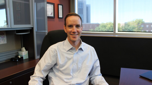A man in a button down shirt sits at an executive desk in an office and smiles.