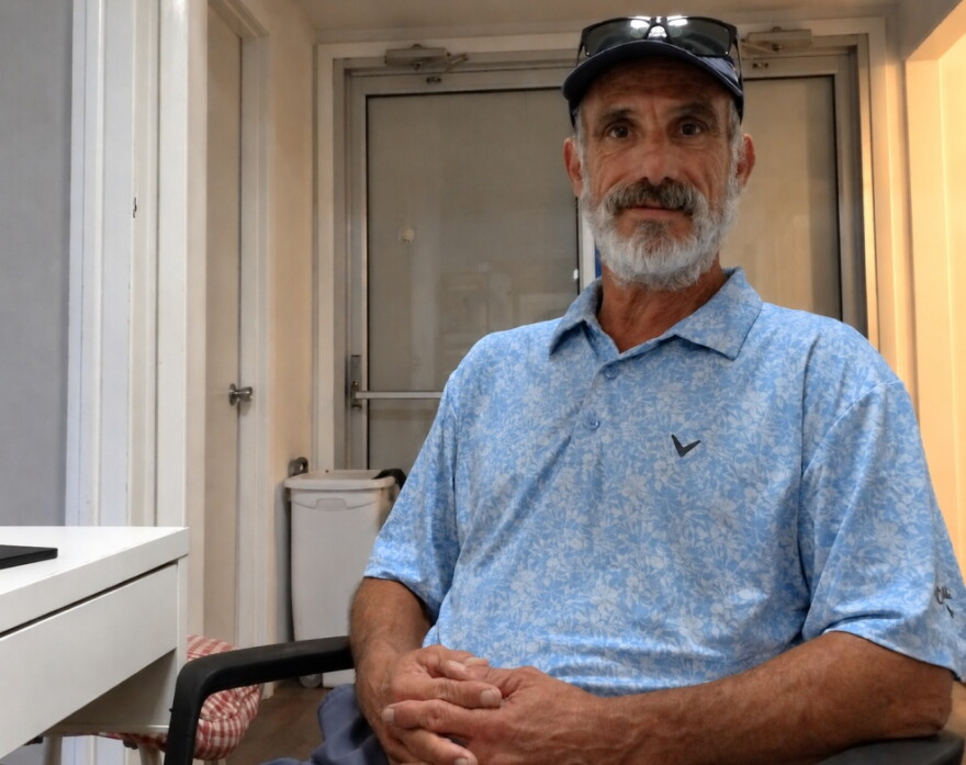 man with full white bears in blueish polo, black baseball cap,  sitting in chair 