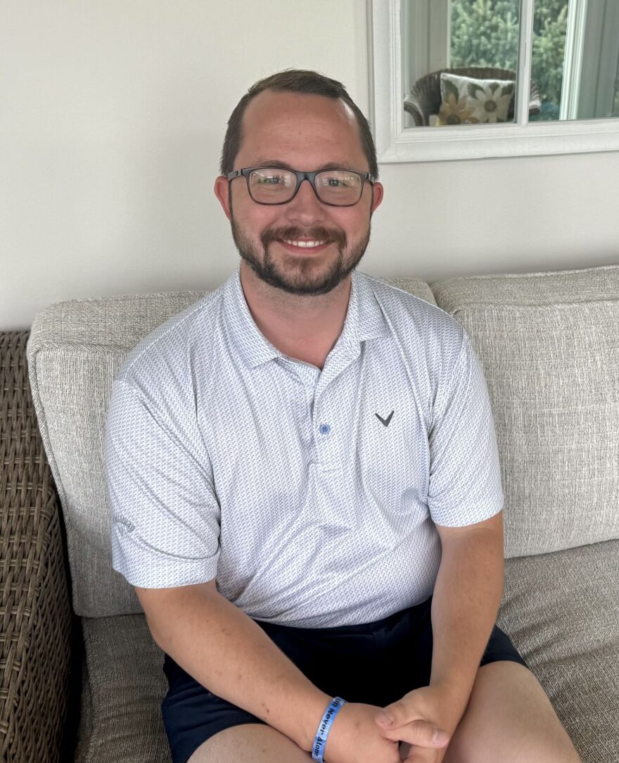 A man is smiling and wearing a white polo shirt with tiny grey markings. He is wearing black shorts. He has brown facial hair and glasses.