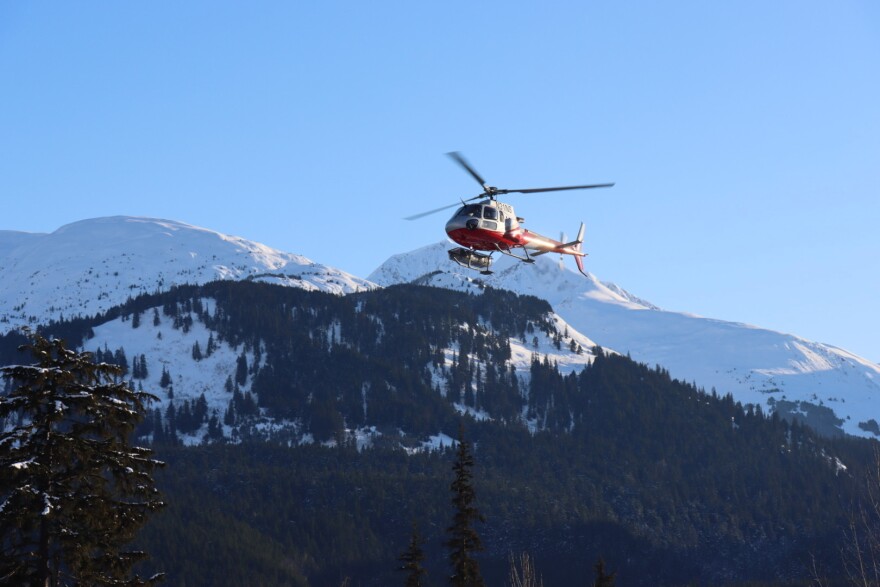 A red-and-white helicopter with a ski basket descending to land with snowy mountains in the background