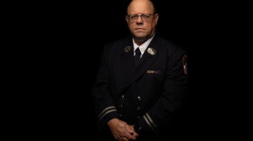 A middle aged man in a firefighter's dress uniform stands in front of a black background with a somber expression.