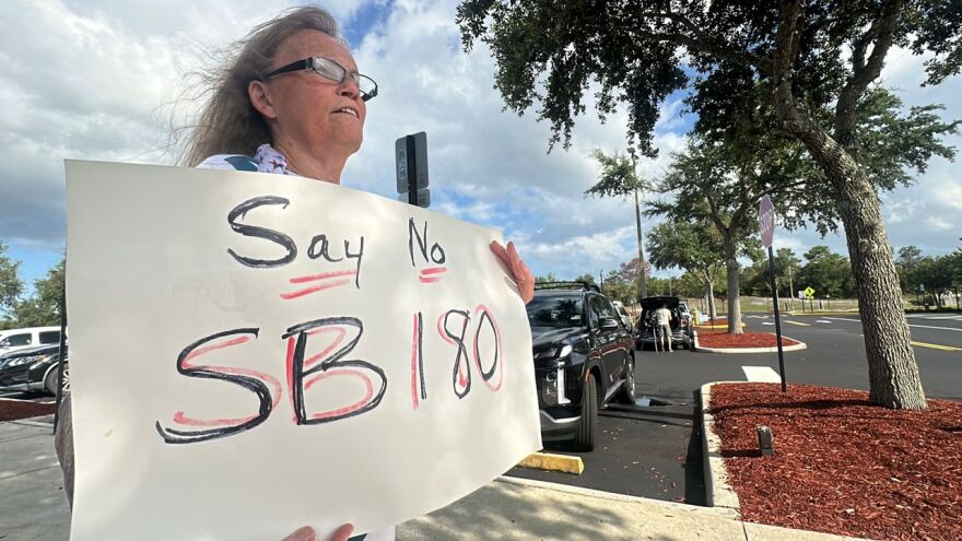 White woman with black glasses holds a sign that reads "Say no SB180"
