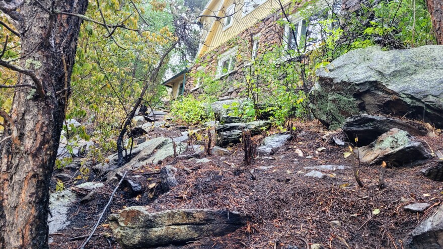 Blackened trees and debris on a hillside come very close to a house with stone and wood siding. 