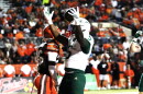 football player in white jersey #5 white helmet and green pants celebrates with both arms raised, football in one hand