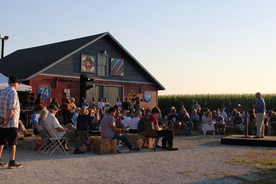 A crowd sits near a barn decorated with road signs, listening to a person speaking on a small stage at sunset. Cornfields and clear skies are in the background.