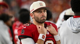 White football player with brown beard holds hands up to his chest wearing a red jersey with other football players blurred out in the photo behind them