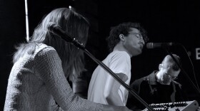 A man sings into a microphone on stage in a small performance space while a woman plays synthesizer in the foreground and another man plays bass in the background.