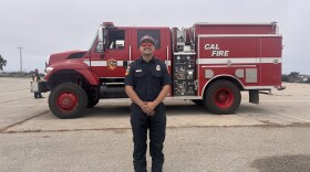 A man in a blue dress firefighter uniform and sunglasses smiles in front of a red Cal Fire truck.