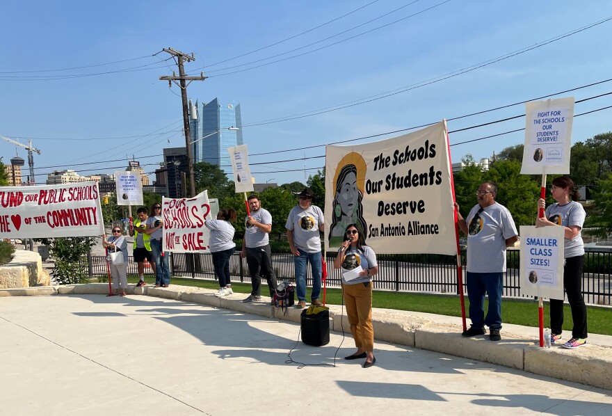 San Antonio Alliance President Alejandra Lopez addresses the crowd with a row of union members holding signs and banners behind her with the downtown skyline in the background.