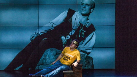 A man on stage in a yellow t-shirt and wearing a gold crown is stretched on a rock, with a projection of a man in Shakespearean style clothing looming behind him.
