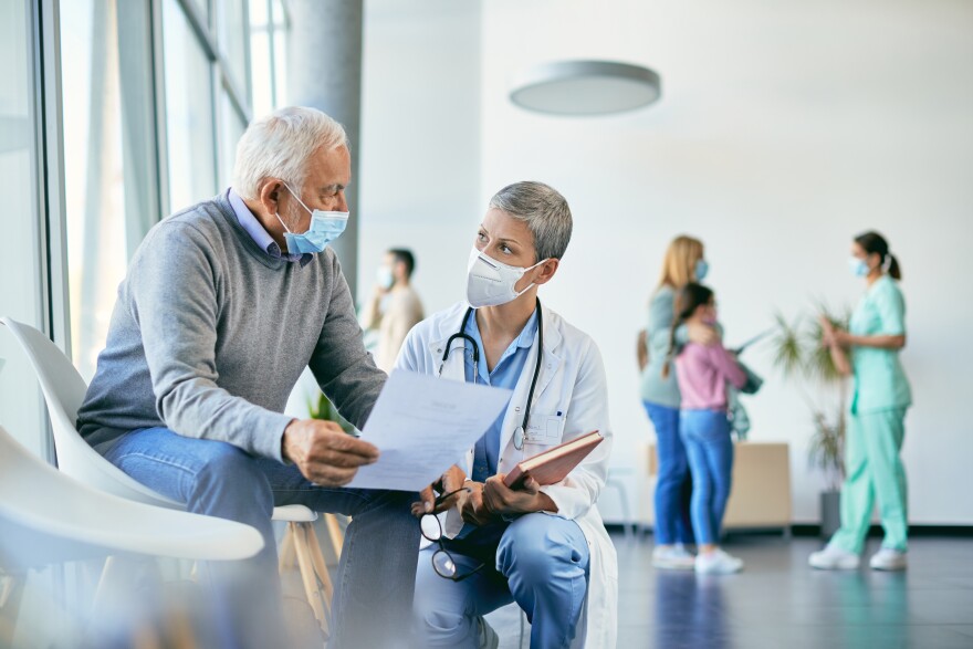 Mature patient communicating with female doctor about medical examination data at the clinic and wearing protective face masks due to COVID-19 pandemic.