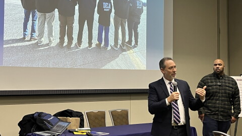Two men stand in a room and projected behind them is an image of DEA officers. 