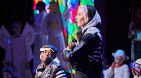 A young woman with a knit scarf wrapped around her head cradles a doll while singing on stage during a theater production.