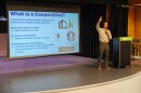 Man on stage holding one arm up in front of screen displaying powerpoint presentation 