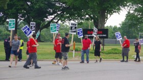 Around ten people stand on the side of the road in front of a chain-link fence and a black sign to the middle right of the image that reads "DAUCH" in red text, below which reads "Shipping & Receiving" in white text with an arrow pointing to the right. The men and women hold different colored signs, but most of them read the same message; "UAW ON STRIKE." One woman's sign to the far right of the image can only be pairly read, it says reads "UNFAIR LABOR PRACTICE" in blue text.