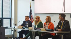 Two men and women seated at a table with microphones and large books in front of them and a projector screen and American flag behind them
