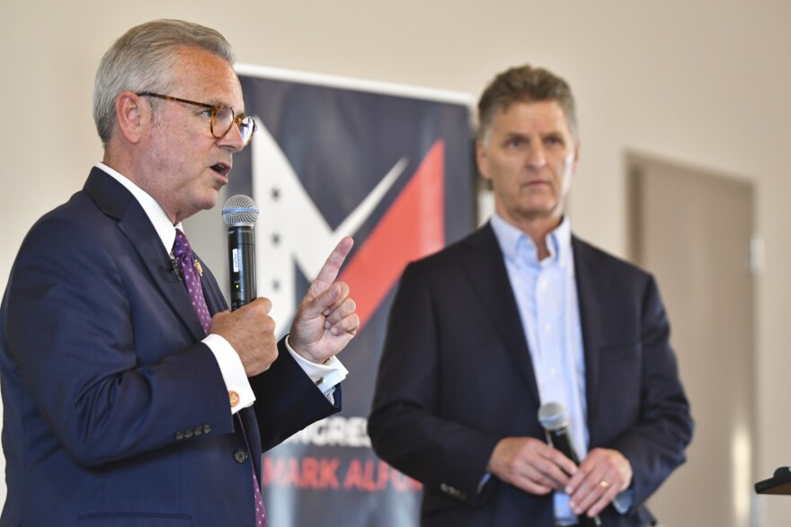 A man wearing a blue suit and tie (Mark Alford) stands indoors (at left) talking on a microphone and gesturing with his left hand. Next to Alford, another man wearing a sport coat (Steve Kraske) is standing and listening to him talk.