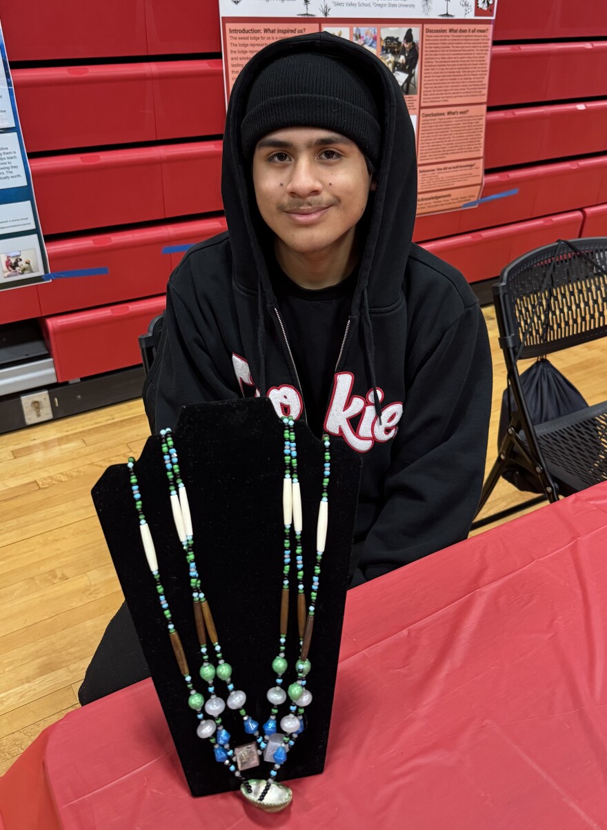Teenager sits at a table behind a display of a necklace made from beads.