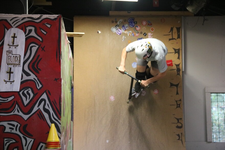 A guy on a scooter wearing a white helmet is riding up the side of a vertical ramp. He is about 9 feet up it. Next to him is a red wall with black and white graffiti art.