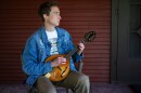 a man sitting on a chair on a porch, playing a new-looking mandolin and gazing off camera