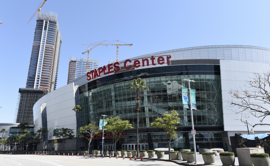 An exterior view of the "STAPLES Center" venue, against a blue sky and with skyscrapers and cranes in the background.