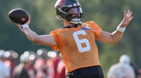 A white man wearing an orange football jersey with the number 6 on it in white font and a dark gray football helmet with the red, white, and black buccaneers flag logo. He has both arms outstretched and is getting ready to throw a football, which is in his right hand.