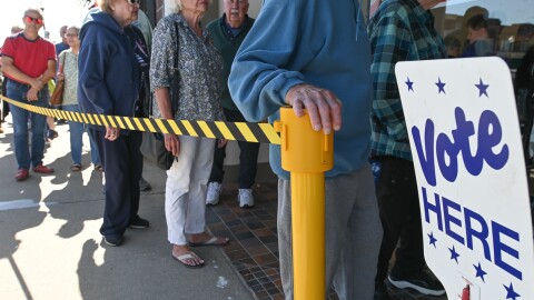 A long line of people stand on a sidewalk behind a yellow tape. In the foreground there is a flag-themed sign that reads "Absentee Voting."