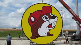 A large circular sign showing Buc-ee’s beaver mascot, a smiling cartoon beaver in a red cap with a white-gloved paw raised, on a yellow background with a dark outline. The sign rests on a metal stand on a concrete lot while a crane holds it up from above. Two workers stand nearby, one sweeping and one by a red bucket, with an overpass and cloudy sky behind them.