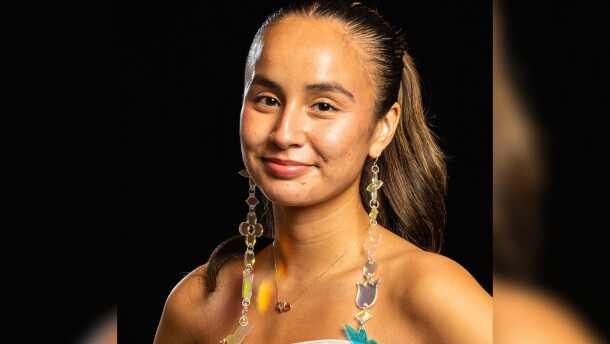 A woman poses against a black background. She is wearing a pink strapless dress and long beaded earrings. 