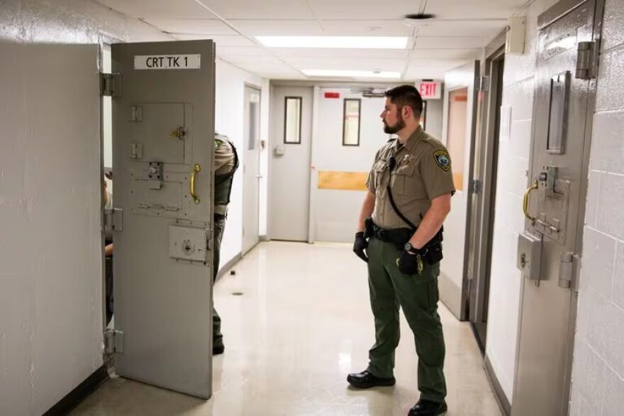 Corrections officer standing in the hallway of a jai. A door to a cell is open. Someone is partially visible through the door.