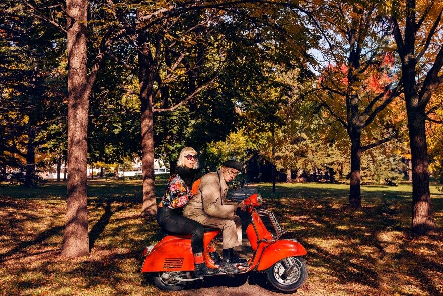 A photo of two people -- Joannie Lafrenière and Gabor Szilasi -- on a red moped with fall foliage and dappled light in the background. 