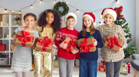 Group of happy smiling diverse children holding Christmas presents tied with beautiful red bows and looking at camera standing in cozy festively decorated living-room during fun party at home