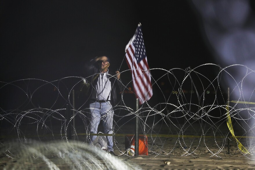 A man holding a United States flag stand on the U.S. side of the border, seen through the concertina wire where the border meets the Pacific Ocean, Thursday, Nov. 15, 2018, from Tijuana, Mexico. 