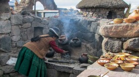 A Quechua woman tends to her clay oven in her outdoor kitchen on the road to the Sillustani archaeological site in Puno, Peru. The stone table is laid with a collection of potatoes and other tubers, as well as homemade cheese and bread.