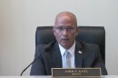 A man in a suit and tie sits at a desk with a nameplate reading "John P. Kahl, Mayor." He faces forward, speaking, with an American flag pin on his lapel and part of an Illinois flag visible on the right.