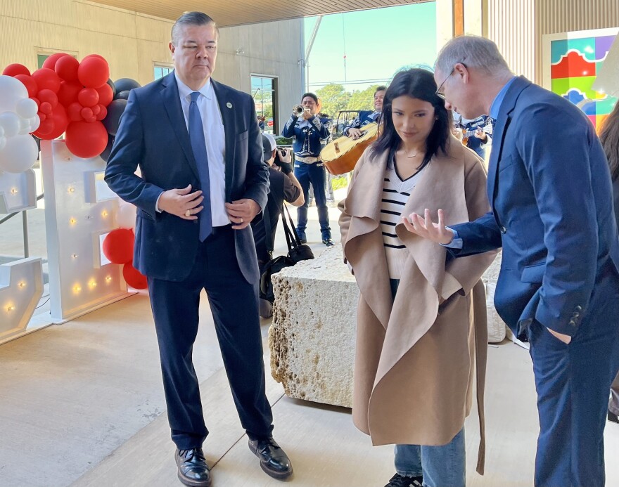 Two men in suits talk to a woman in a tan coat as a mariachi band plays in the background.