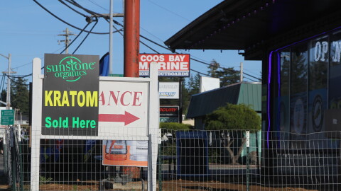 A strip mall during the day. A sign reads "Kratom Sold Here" outside of a smoke shop in a strip mall.