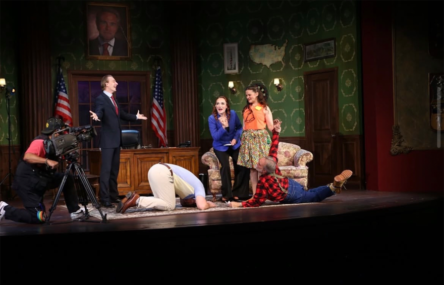 Six people appear on a set made to look like a governor's office. Two men are on the floor, two women stand, one man stands with his hands stretched out to either side of him, and another man kneels behind a camera. 