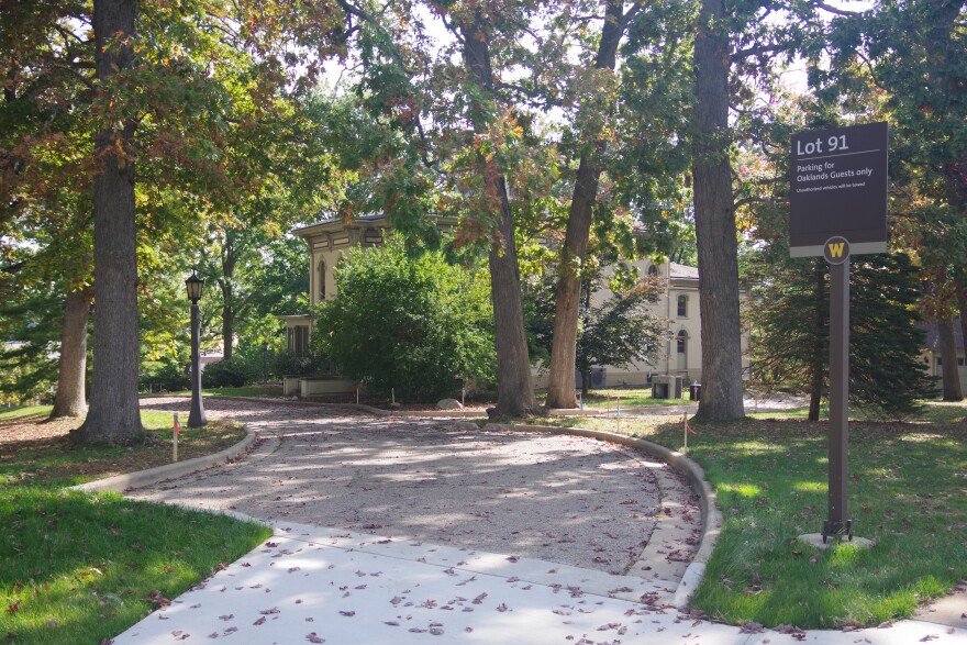 An off-white two-story building is obscured by trees and bushes, which populated much of the image. Most of the trees stand taller than the image can capture, with branches dangling down into the top of the frame. A road cuts through the trees, with small wooden stakes alongside it. A brown metal sign on the right designates the area as "Lot 91," describing what permits are valid for parking. 