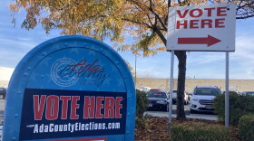 In the foreground, a blue ballot box  with the words "Vote Here. AdaCountyElections.com" written in the front of it. In the background, a parking lot with fall leaves on the ground, a tree and a tall white sign with the words "Vote Here" and an arrow pointing to the right. 