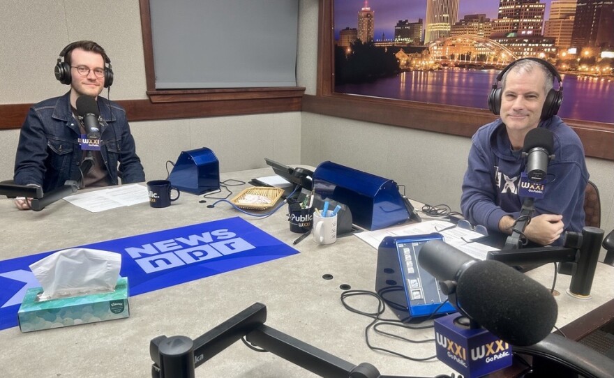 Two smiling men wearing headphones sit at a table in a radio talk show: the man at left has short brown hair and is wearing glasses, a brown shirt, and a denim jacket; the man at right has short dark hair and is wearing a navy blue hoodie with "WXXI" in white lettering