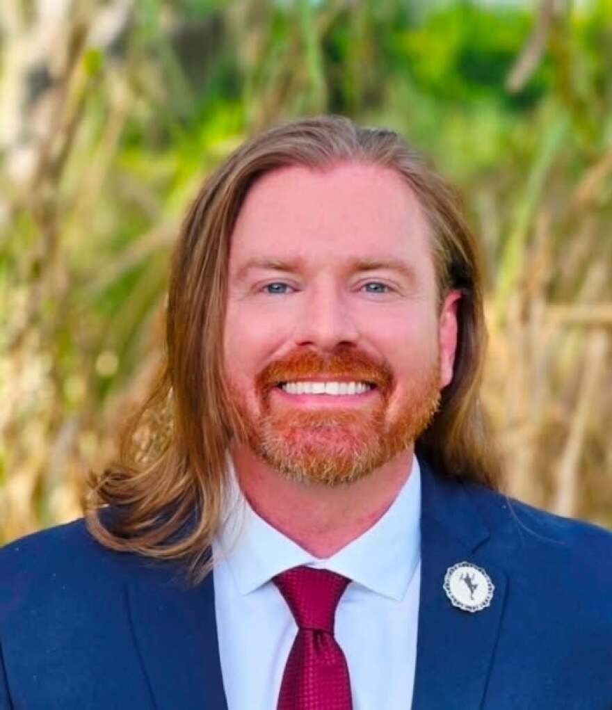 Man with shoulder-length reddish hair and beard wearing a blue suit and smiling into the camera