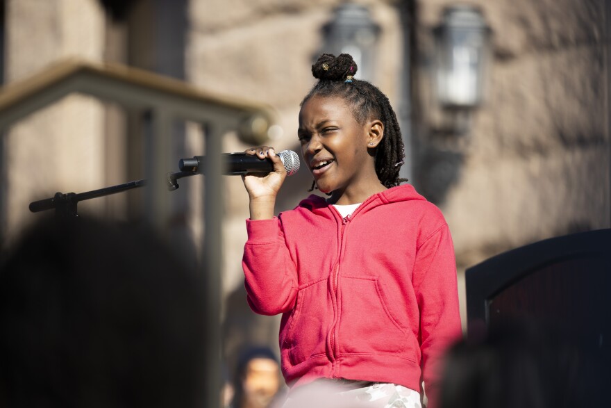 A young girl dressed in a red jacket stands in front of the Texas Capitol. She holds a microphone and is delivering a speech to the audience.