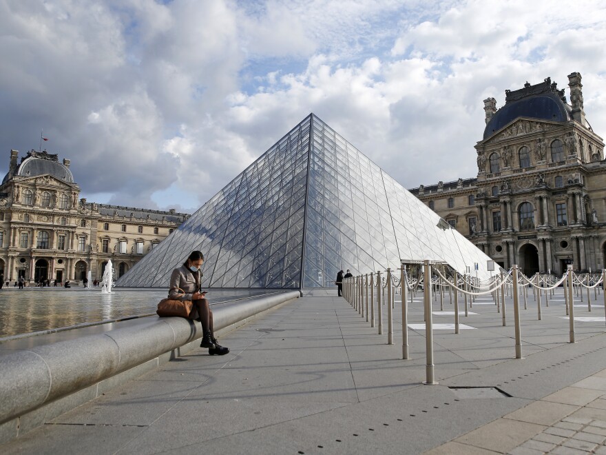 French President Emmanuel Macron is ordering nightly curfews for Paris and other large cities, hoping to control the coronavirus. A woman wears a protective face mask as she sits in the Louvre Museum's courtyard Wednesday in Paris.