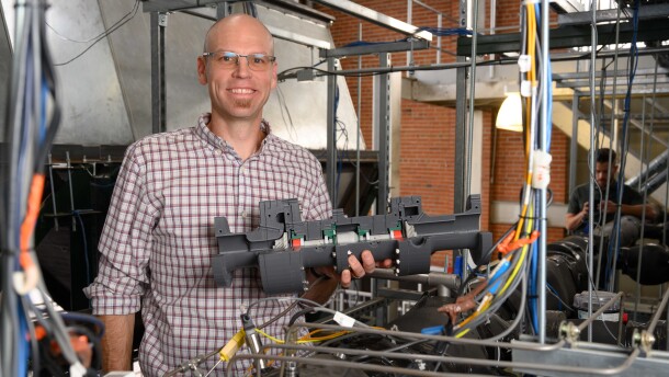 A man smiles while holding up a piece of machinery, while surrounded by wires in some sort of lab or factory