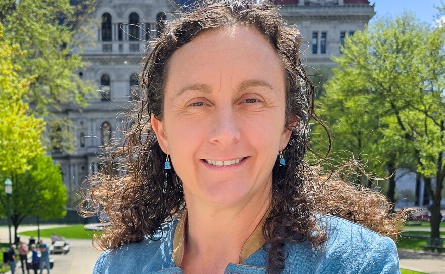 A woman with long dark hair stands outside wearing a light blue blazer over a black and white patterned top.