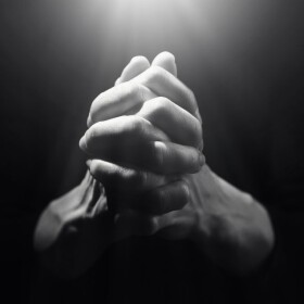 Hands clasped together in prayer against dark background with light beaming down